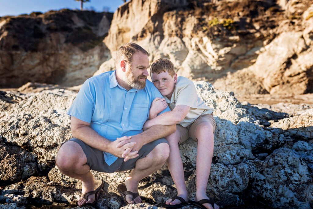 A father and son sit close together on rocky cliffs by the ocean, sharing a quiet, affectionate moment under golden evening light.