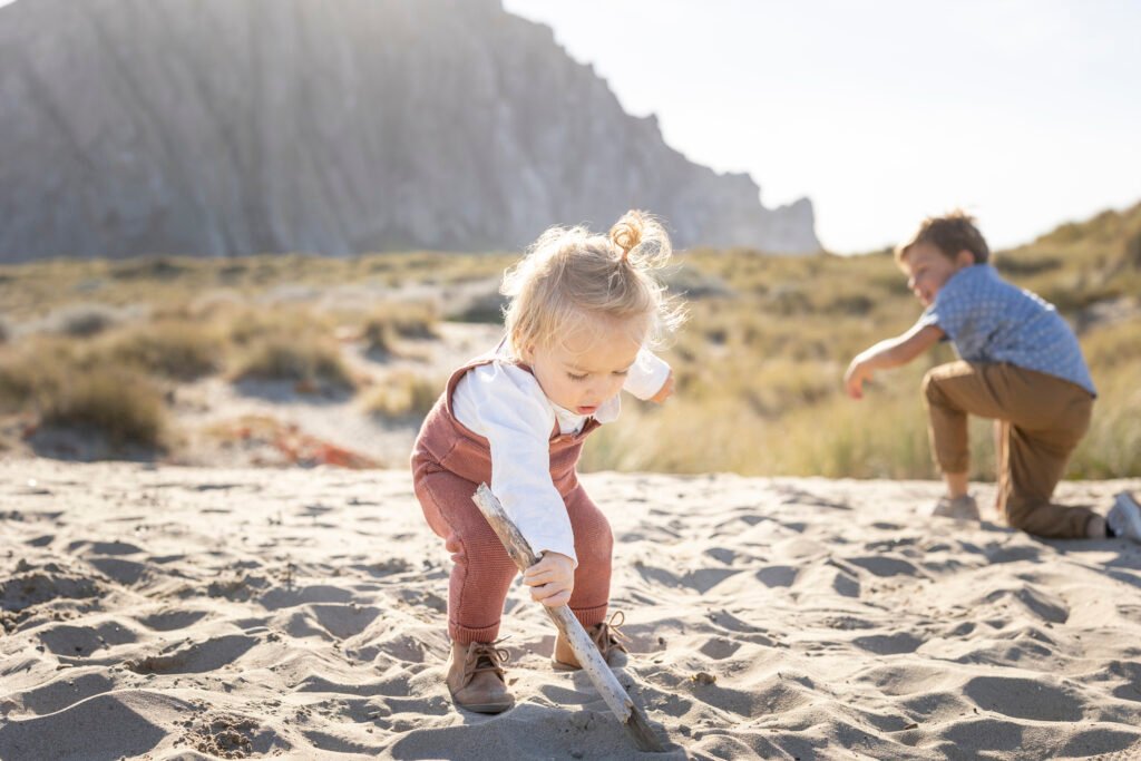 A toddler plays in the sand holding a stick, while a young boy kneels nearby, both surrounded by beach grasses with Morro Rock in the distance.